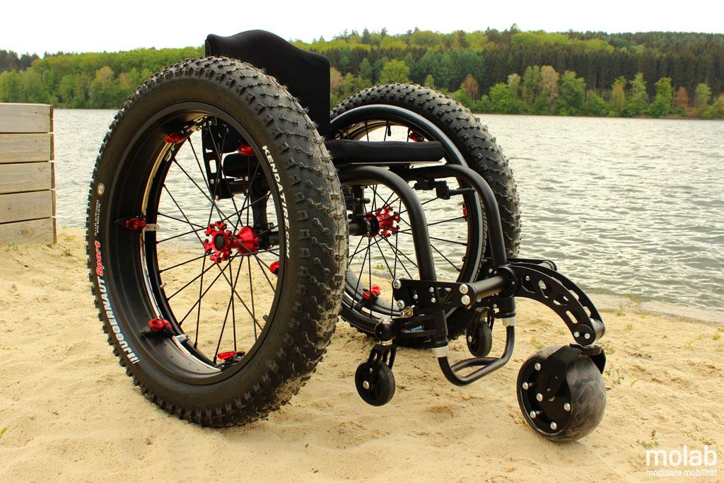 Straponwheelz mit Smartwheel montiert an einem leeren Rollstuhl stehend im Sand an einem See
